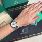 Wristwatch on a left wrist: silver Rolex with white dial and diamond hour markers, beside green Rolex box and Privilegia card.