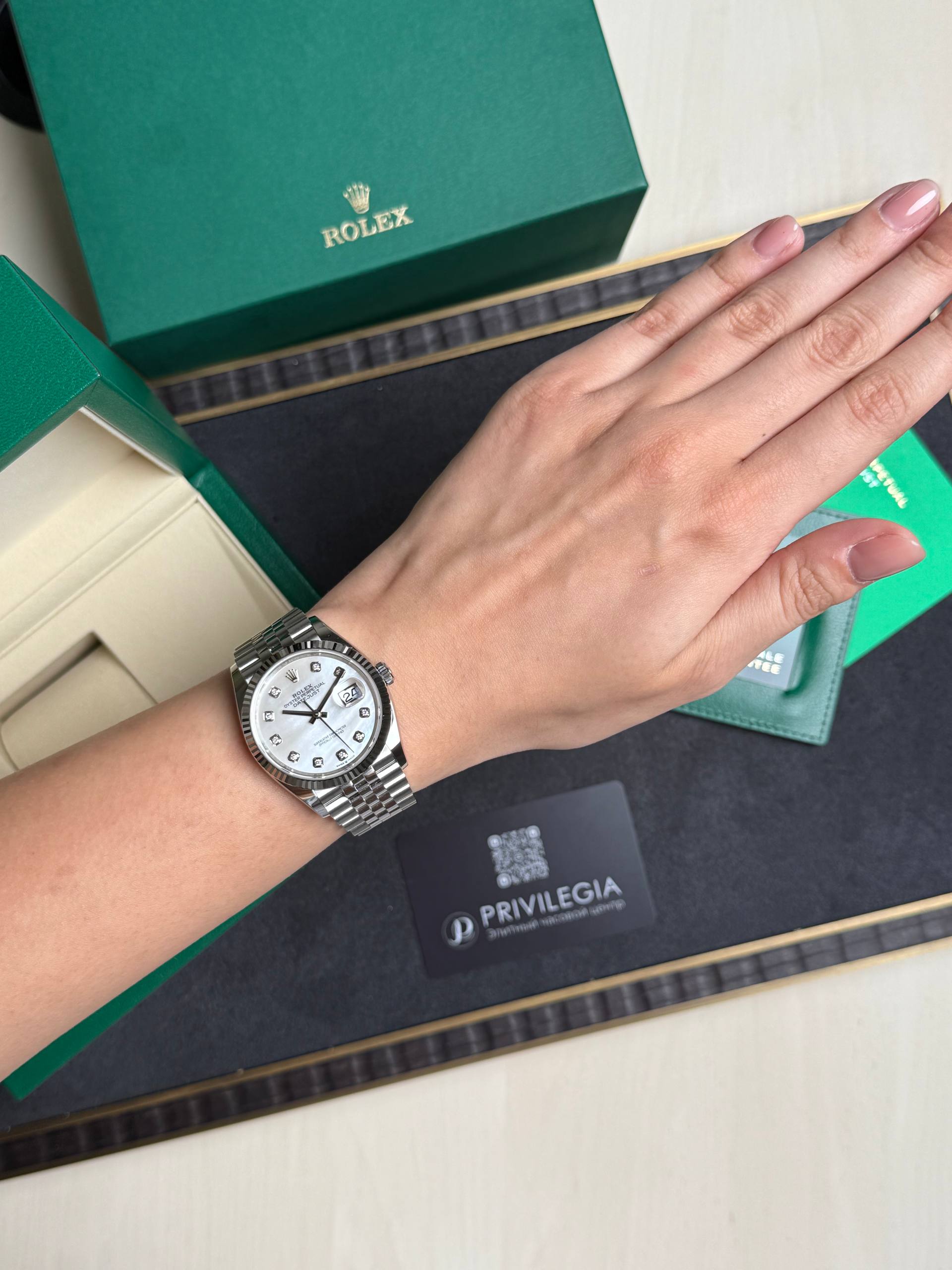 Wristwatch on a left wrist: silver Rolex with white dial and diamond hour markers, beside green Rolex box and Privilegia card.