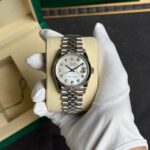 Rolex Datejust in a green box, silver bracelet, diamond hour markers, held by gloved hand.