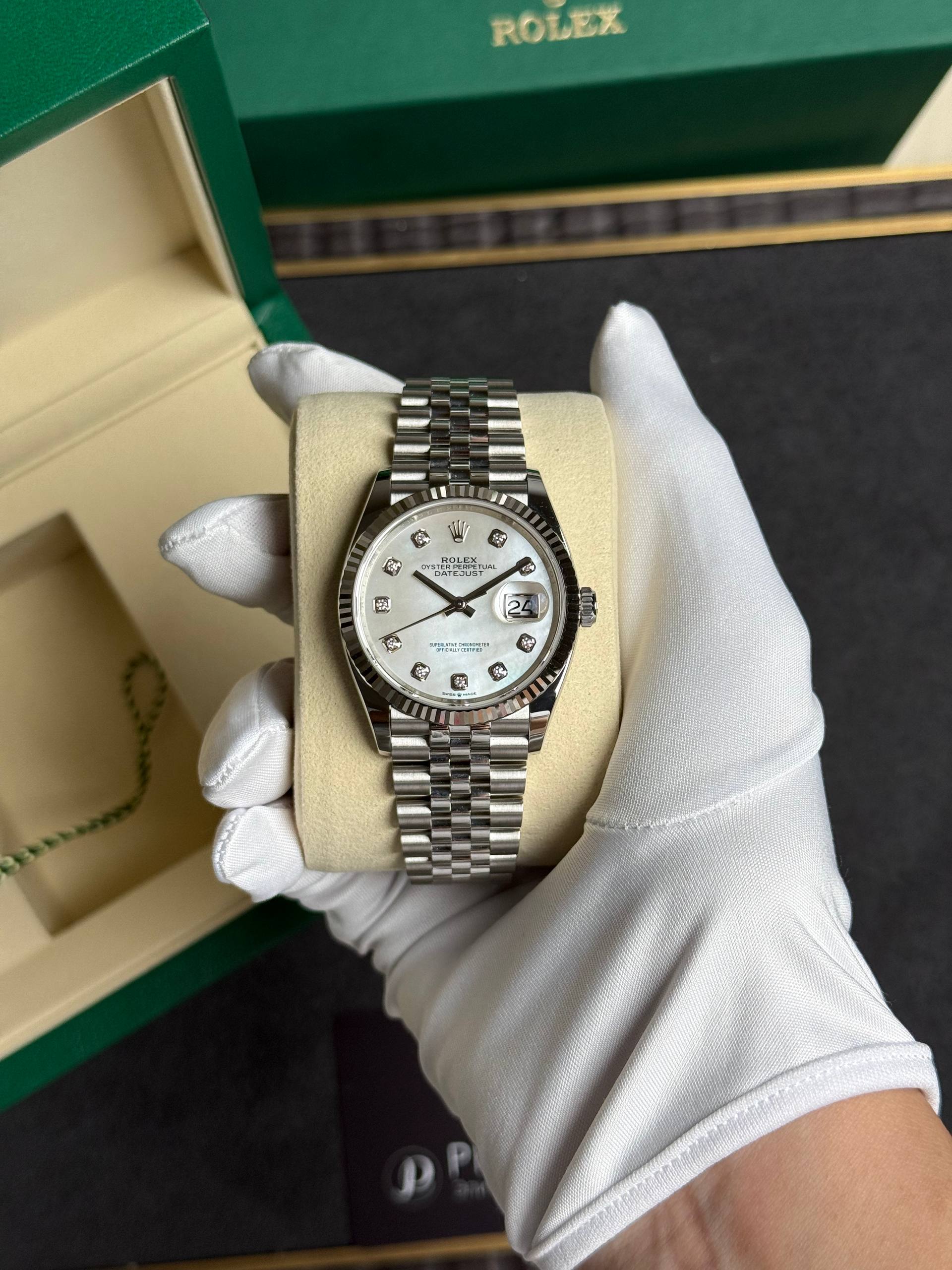 Rolex Datejust in a green box, silver bracelet, diamond hour markers, held by gloved hand.