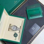 Open green Rolex box revealing a silver Rolex wristwatch in a cream interior with a green seal/pendant below it.