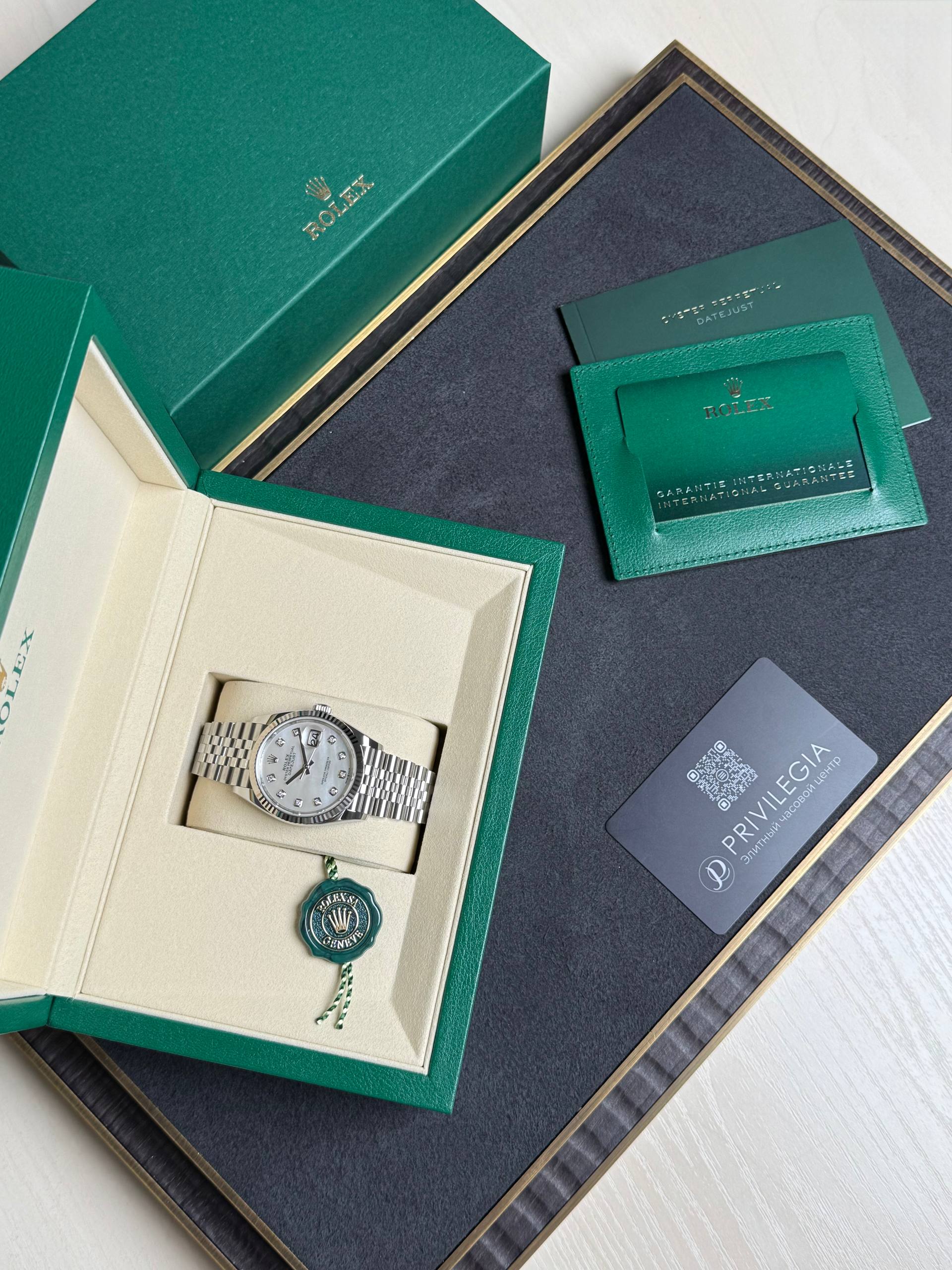 Open green Rolex box revealing a silver Rolex wristwatch in a cream interior with a green seal/pendant below it.