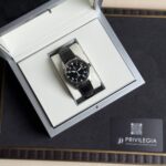 Black-dial wristwatch with white numerals in a padded silver presentation box with a black leather strap and cream interior.