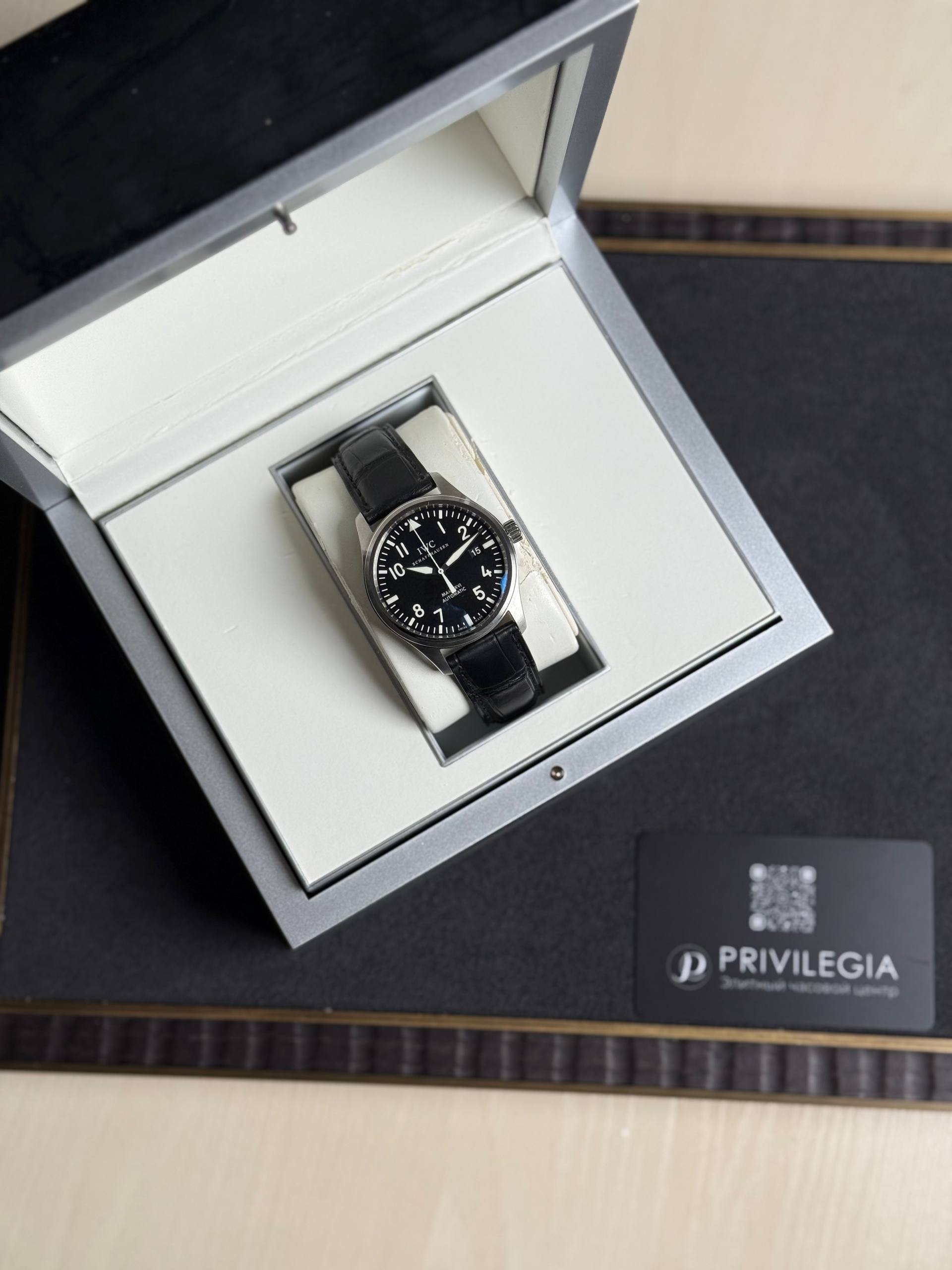 Black-dial wristwatch with white numerals in a padded silver presentation box with a black leather strap and cream interior.