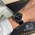 Close-up of a black-dial IWC Pilot watch on a wrist with a white shirt sleeve visible.