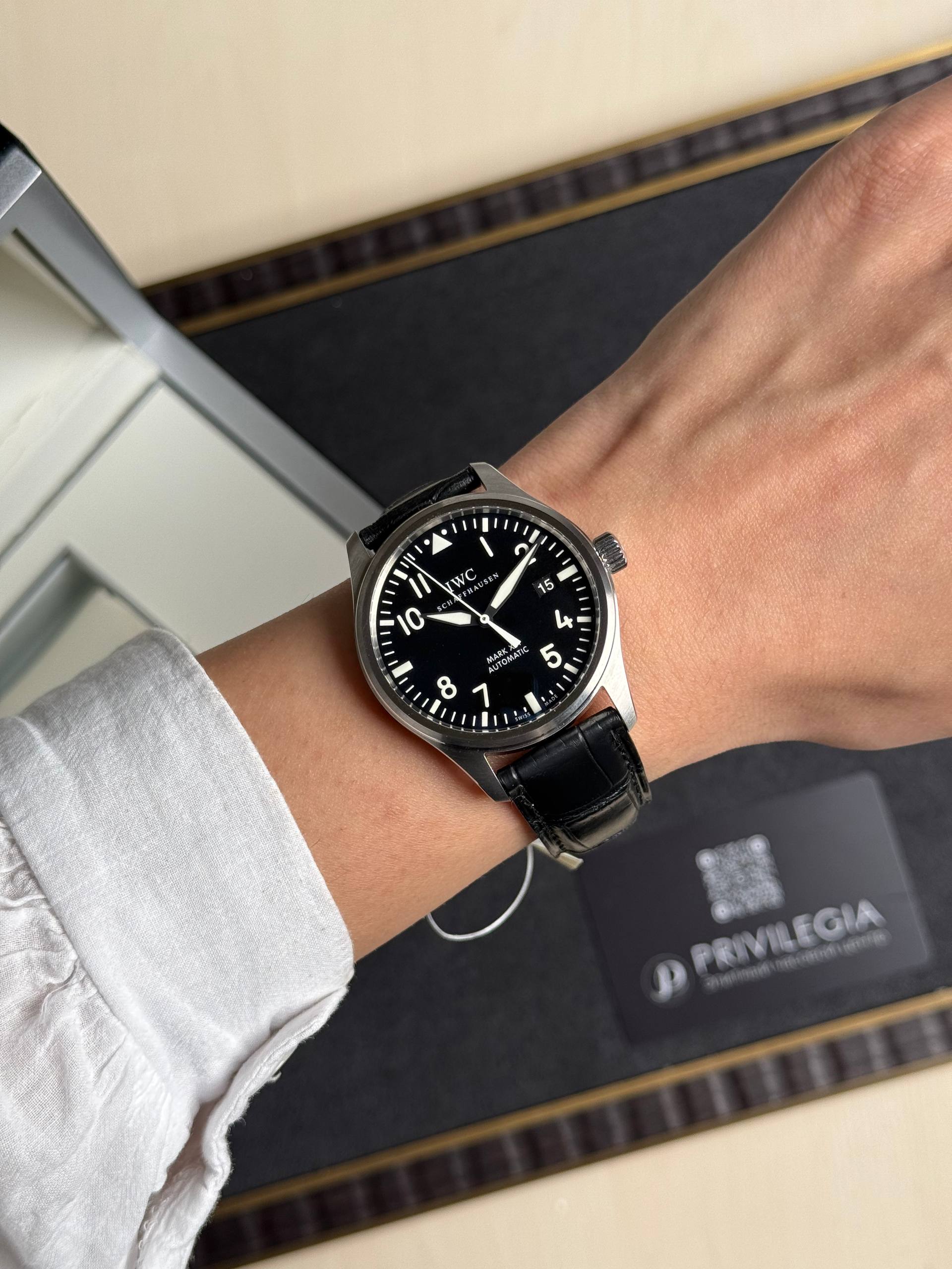Close-up of a black-dial IWC Pilot watch on a wrist with a white shirt sleeve visible.
