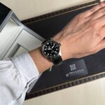 Wrist wearing a black IWC pilot watch with a leather strap resting near a watch presentation box.