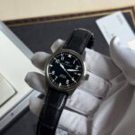 Close-up of an IWC pilot watch with a black dial and leather strap held by a gloved hand.