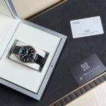 IWC wristwatch in a silver presentation box with black strap on a white cushion, positioned on a dark mat next to a card and QR code.