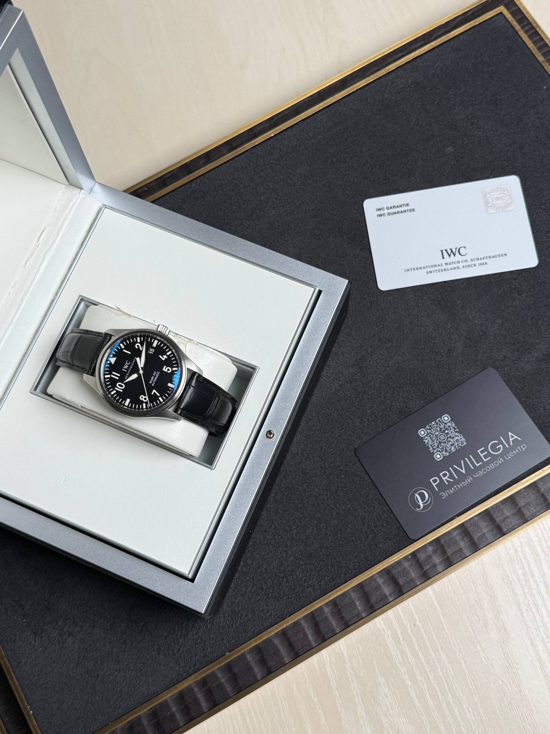 IWC wristwatch in a silver presentation box with black strap on a white cushion, positioned on a dark mat next to a card and QR code.