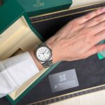 Silver Rolex wristwatch worn on the left wrist, resting on an open green Rolex box and presentation tray.
