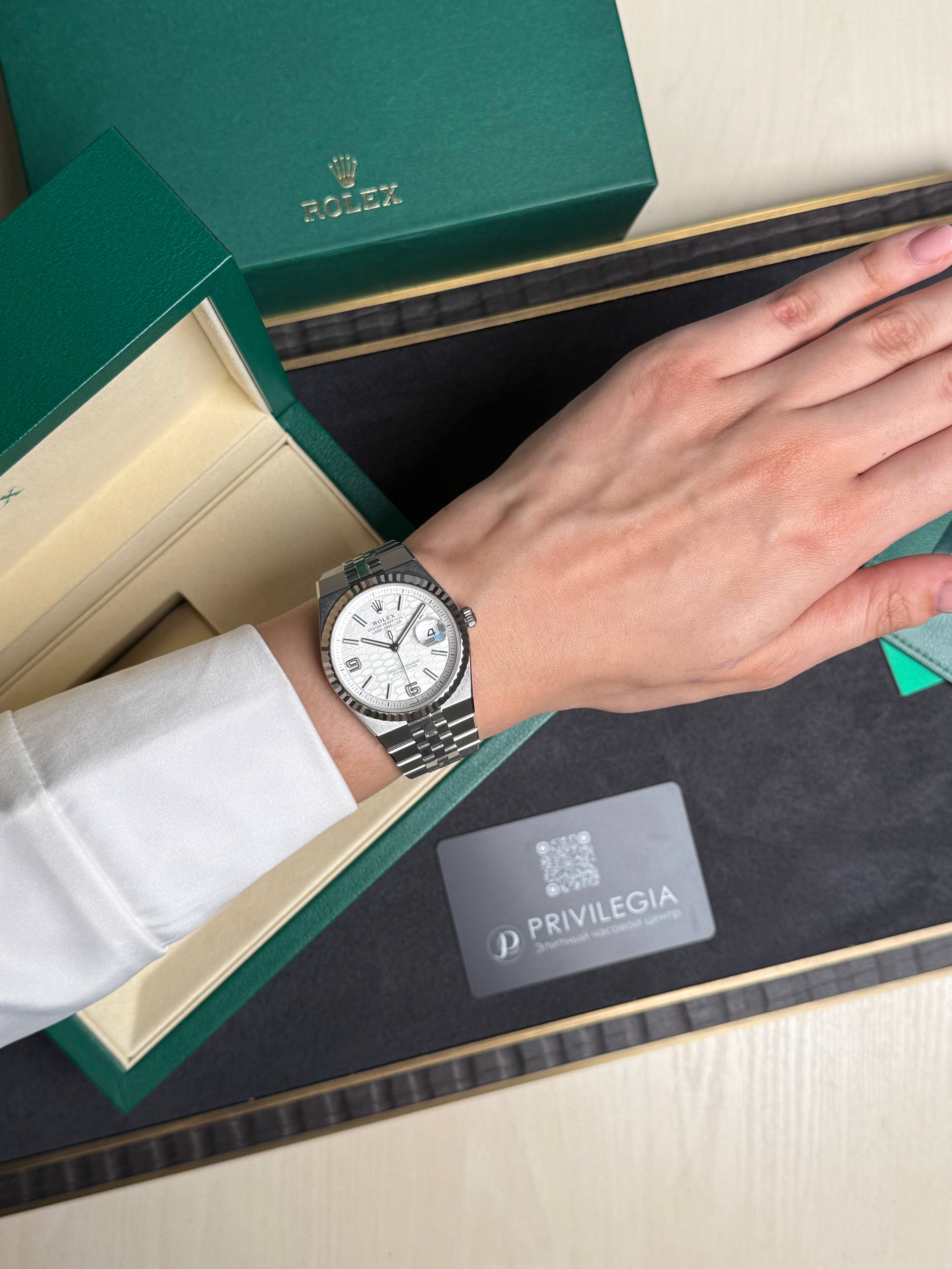 Silver Rolex wristwatch worn on the left wrist, resting on an open green Rolex box and presentation tray.