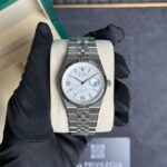 Rolex stainless steel watch with a white patterned dial and fluted bezel on a beige cushion, held by a black-gloved hand in front of a green box.