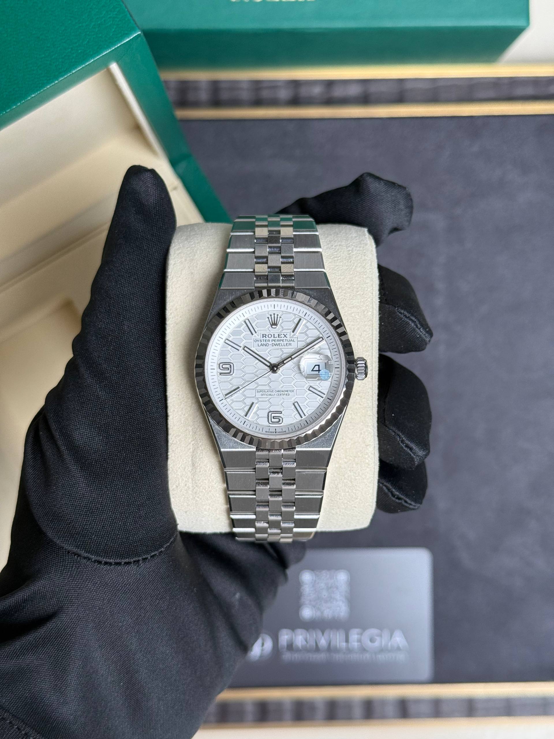 Rolex stainless steel watch with a white patterned dial and fluted bezel on a beige cushion, held by a black-gloved hand in front of a green box.