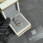 Open jewelry box displaying a diamond-encrusted circular jewelry piece inside a grey velvet interior, held by a gloved hand.
