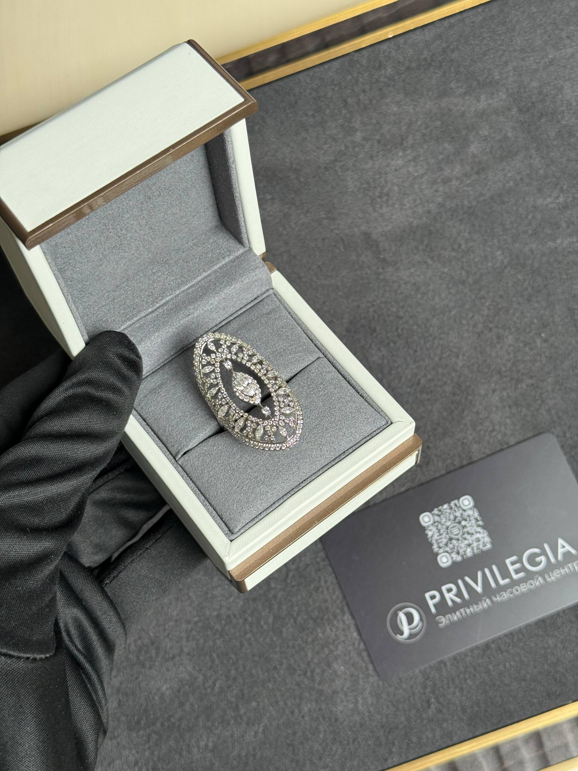 Open jewelry box displaying a diamond-encrusted circular jewelry piece inside a grey velvet interior, held by a gloved hand.