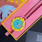 Colorful wristwatch in a pink and yellow box with a turquoise dial and yellow bezel, partially tucked open in packaging.