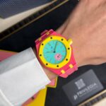 Bright pink wristwatch with a yellow bezel and turquoise dial on a person’s wrist