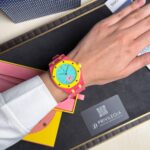 Bright pink Swatch watch with a yellow bezel and turquoise dial on a left wrist, placed over a desk with brand materials nearby.