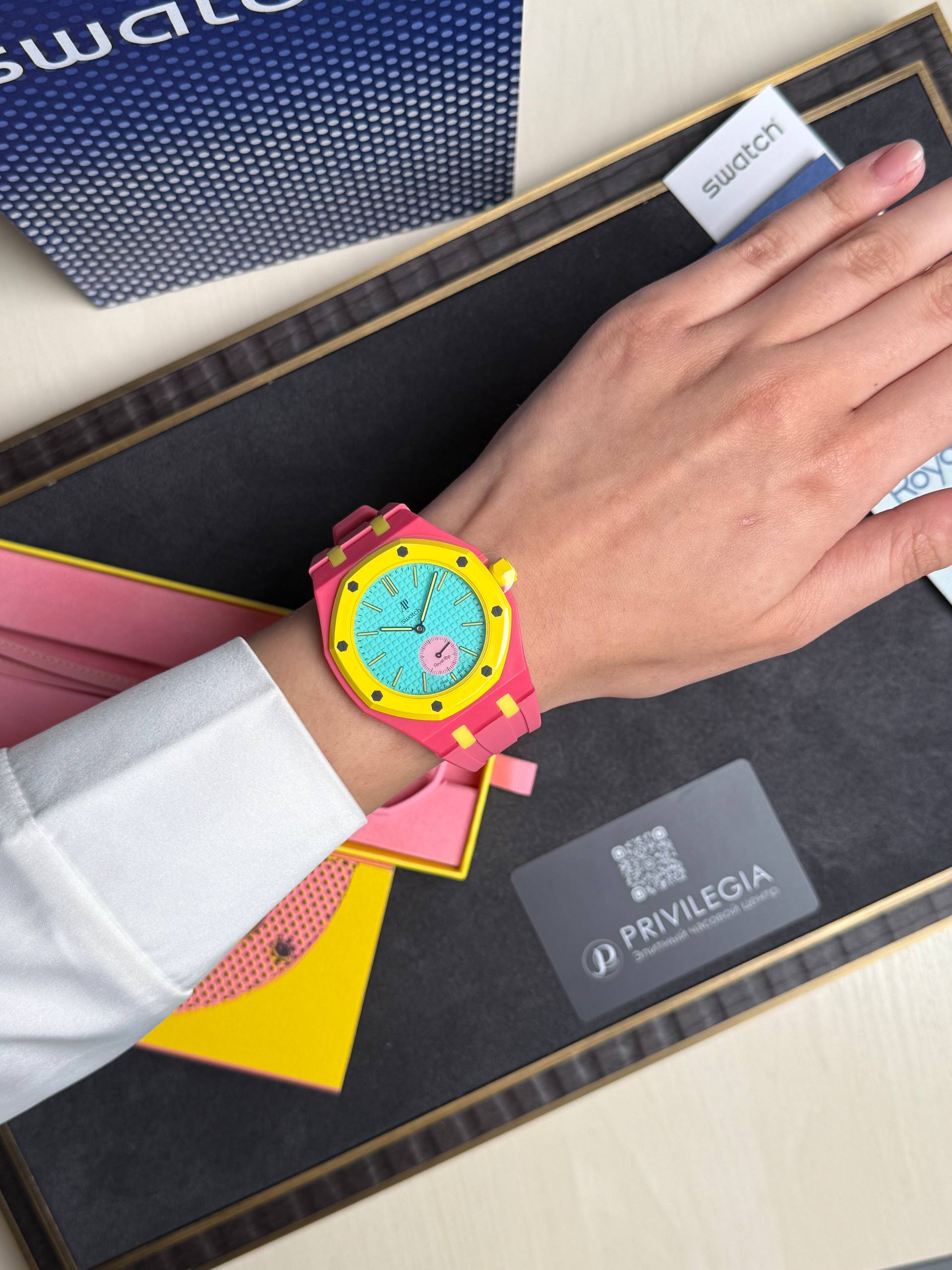 Bright pink Swatch watch with a yellow bezel and turquoise dial on a left wrist, placed over a desk with brand materials nearby.