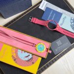 Bright pink Swatch watch with turquoise face resting in a yellow box among colorful packaging and business cards on a dark board surface.