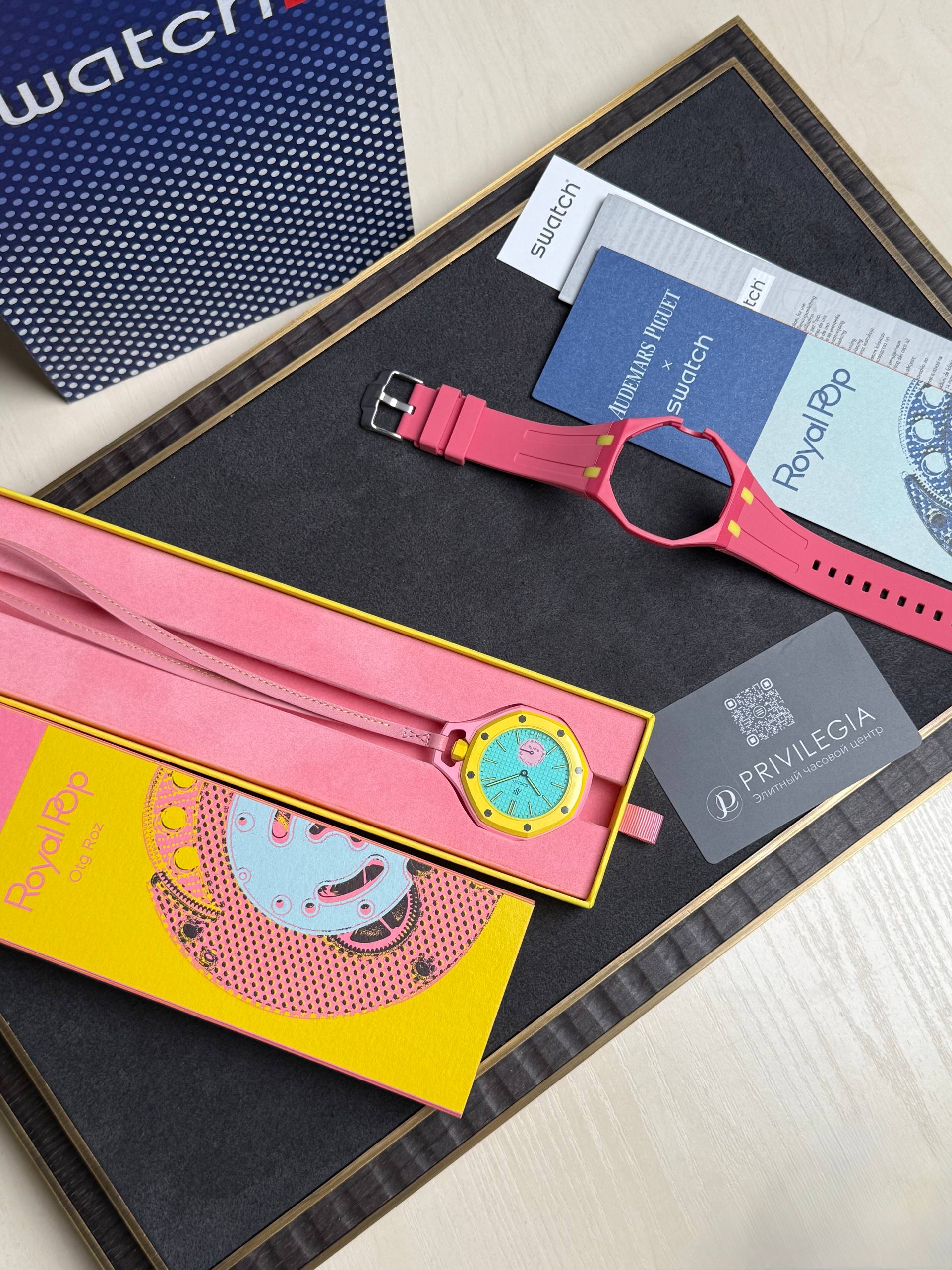Bright pink Swatch watch with turquoise face resting in a yellow box among colorful packaging and business cards on a dark board surface.