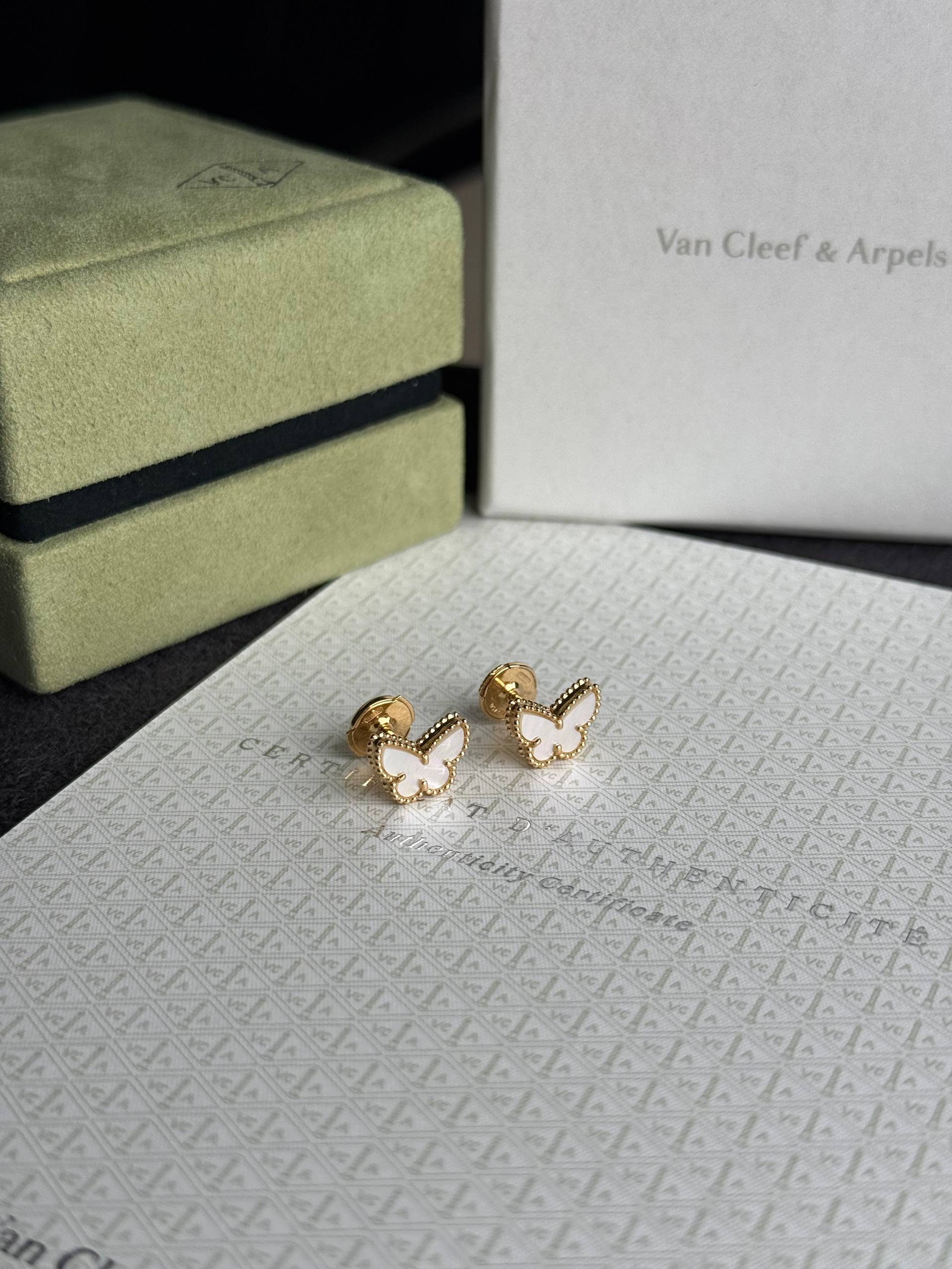 Gold butterfly-shaped earrings with white enamel centers resting on a patterned surface, with a pale green velvet box and a Van Cleef & Arpels box in the background.