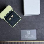 Green velvet jewelry case open to reveal gold butterfly earrings beside a gray Van Cleef & Arpels box and a Privilegia card with a QR code.