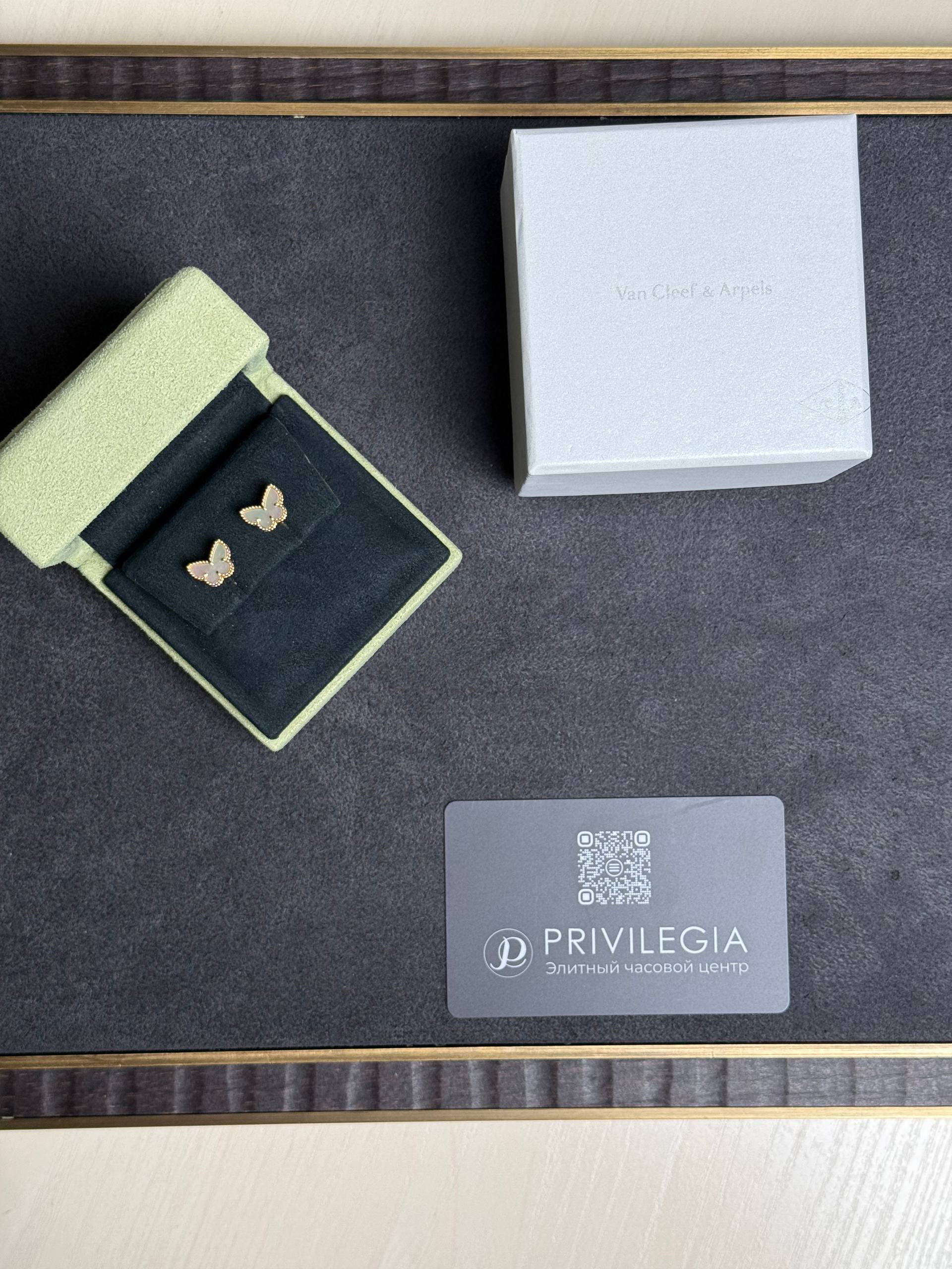 Green velvet jewelry case open to reveal gold butterfly earrings beside a gray Van Cleef & Arpels box and a Privilegia card with a QR code.