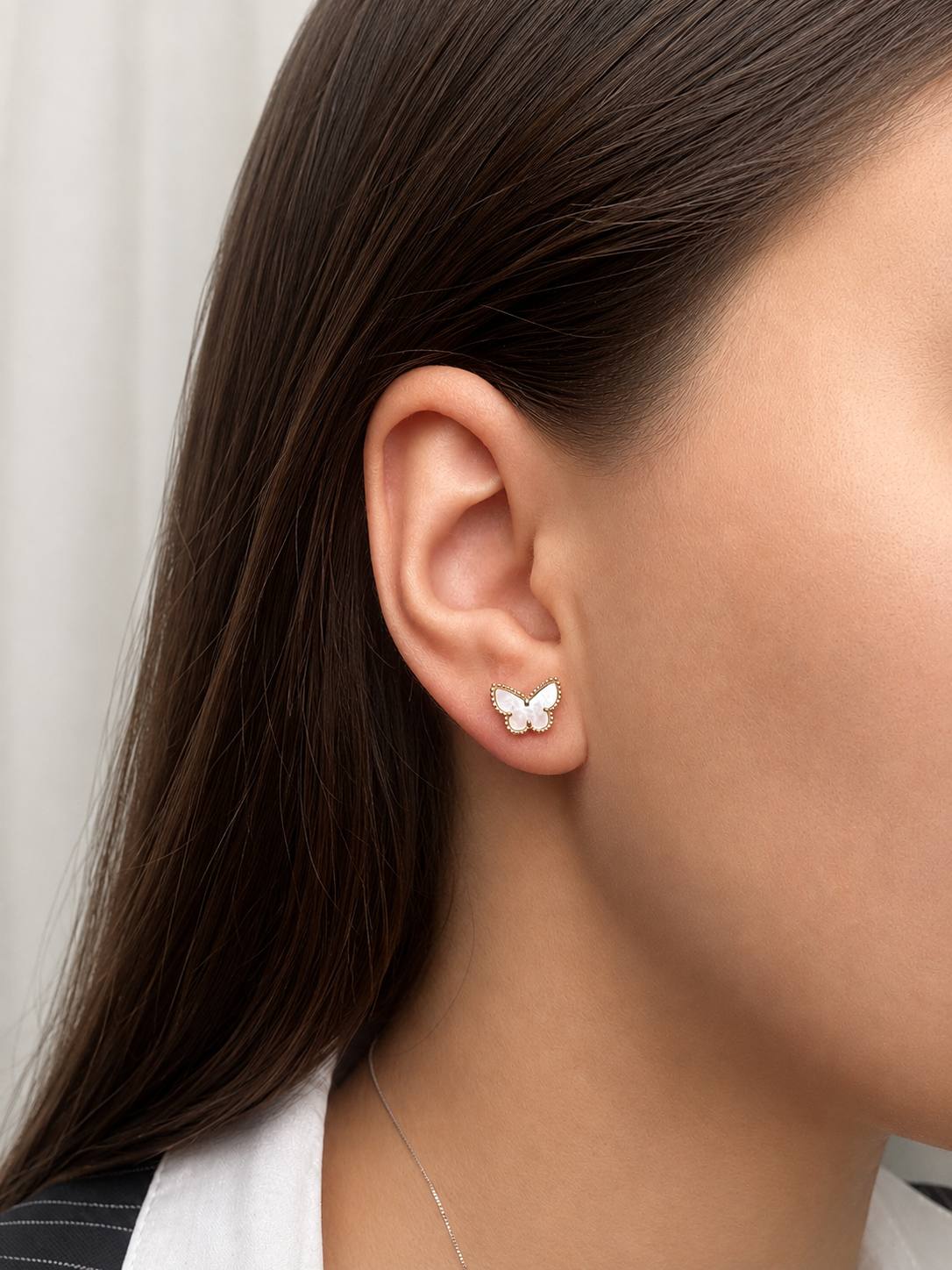 Butterfly-shaped stud earring in white with a gold beaded edge on a model's earlobe, brunette hair tucked behind the ear