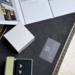 Flat lay of luxury jewelry packaging: white boxes, a gray card with a QR code, and light green and black velvet pouches containing butterfly-shaped charms on a dark surface.