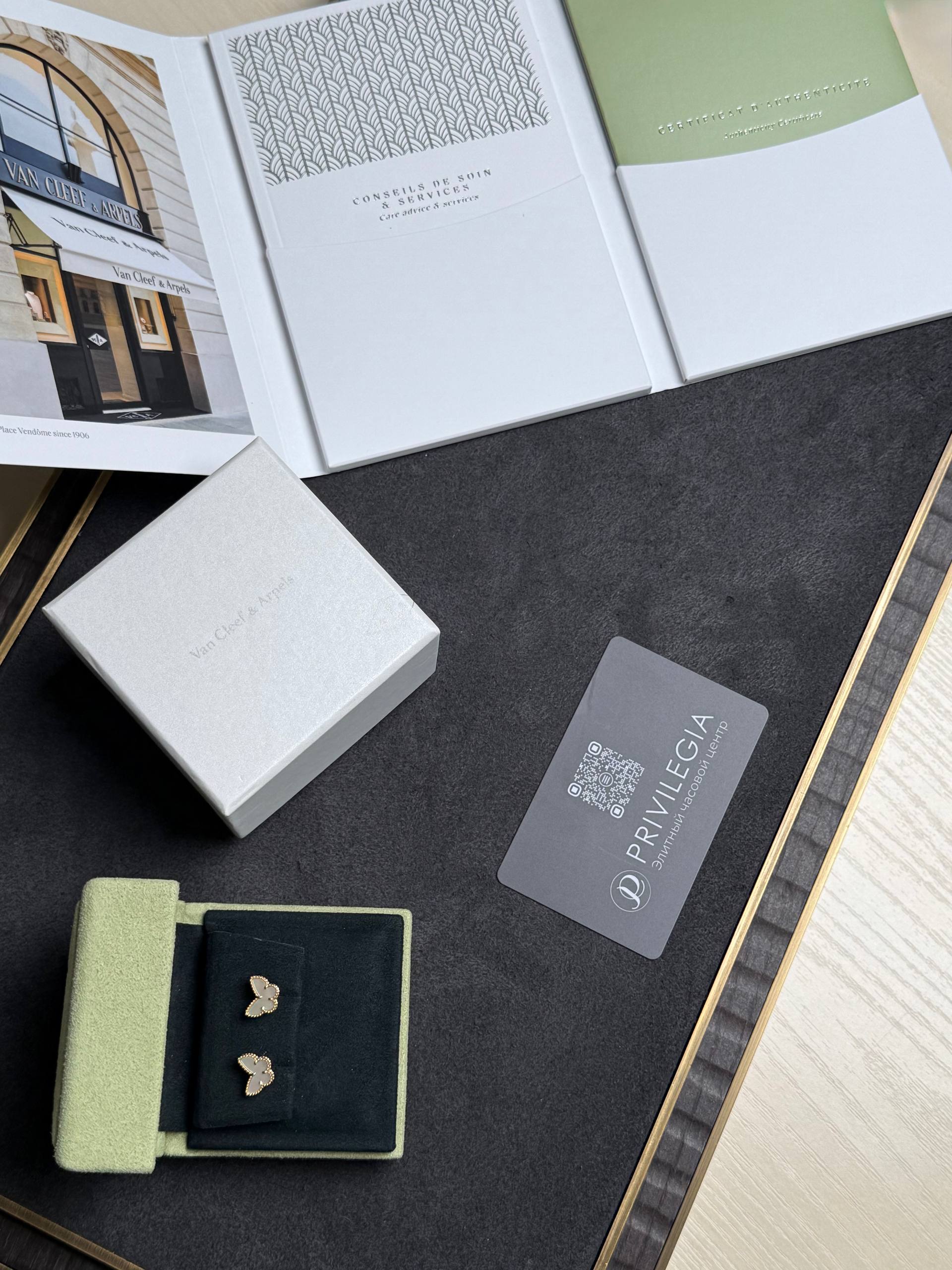 Flat lay of luxury jewelry packaging: white boxes, a gray card with a QR code, and light green and black velvet pouches containing butterfly-shaped charms on a dark surface.
