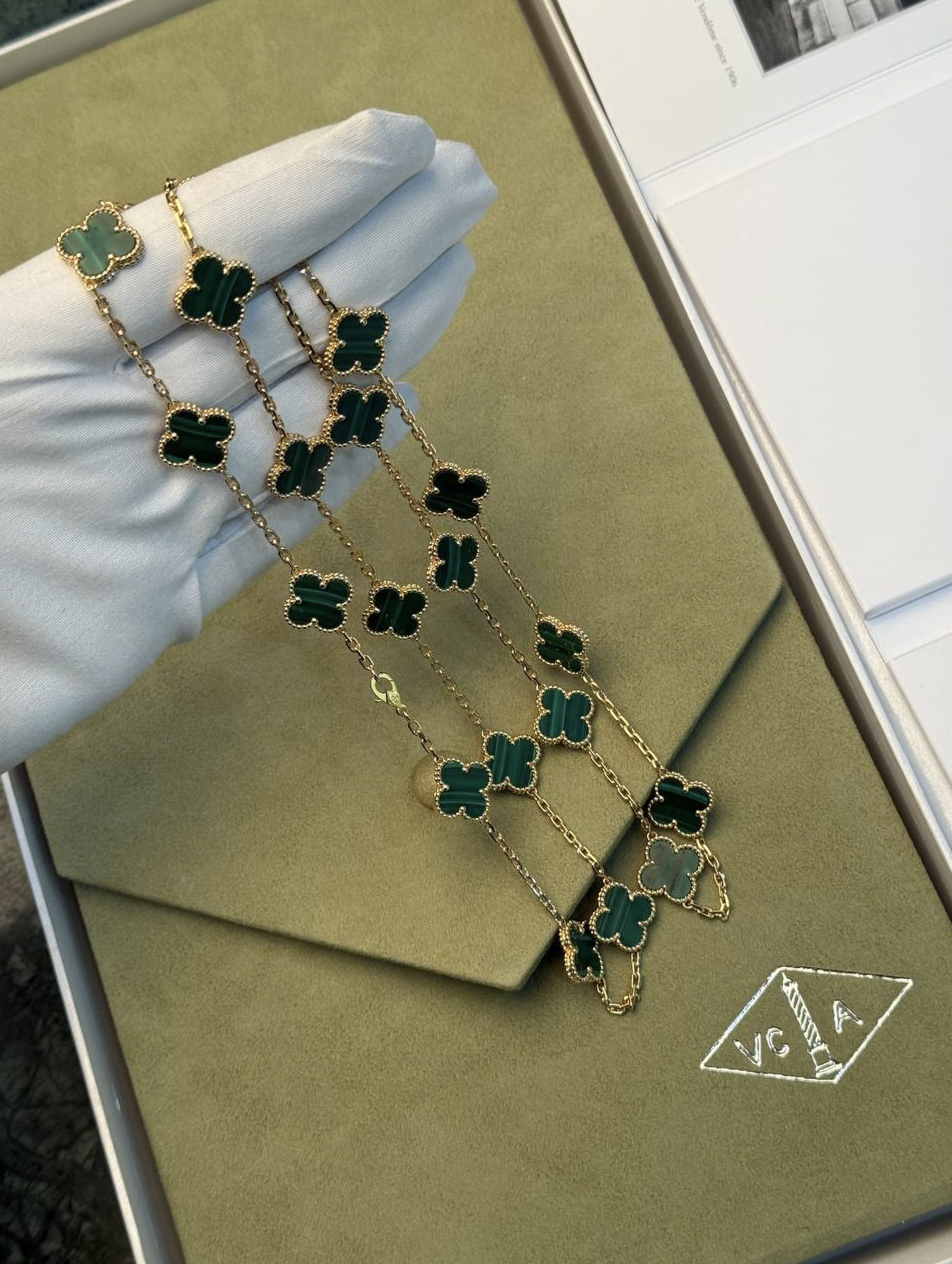 Gold chain necklace with emerald green clover pendants outlined in gold, resting on a green velvet jewelry box.