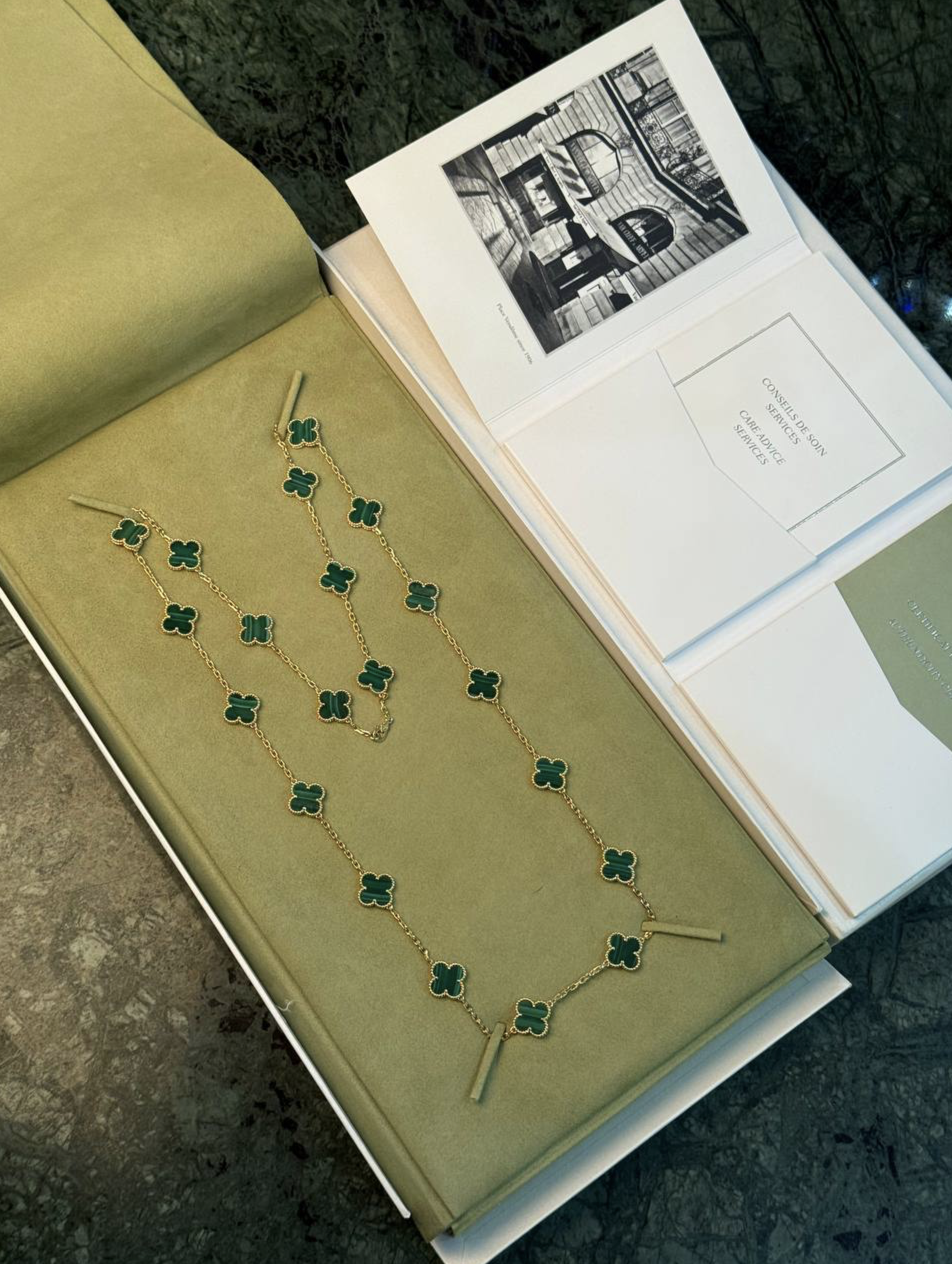 Gold chain necklace with green clover charms arranged in a pale green jewelry box with an open book nearby.