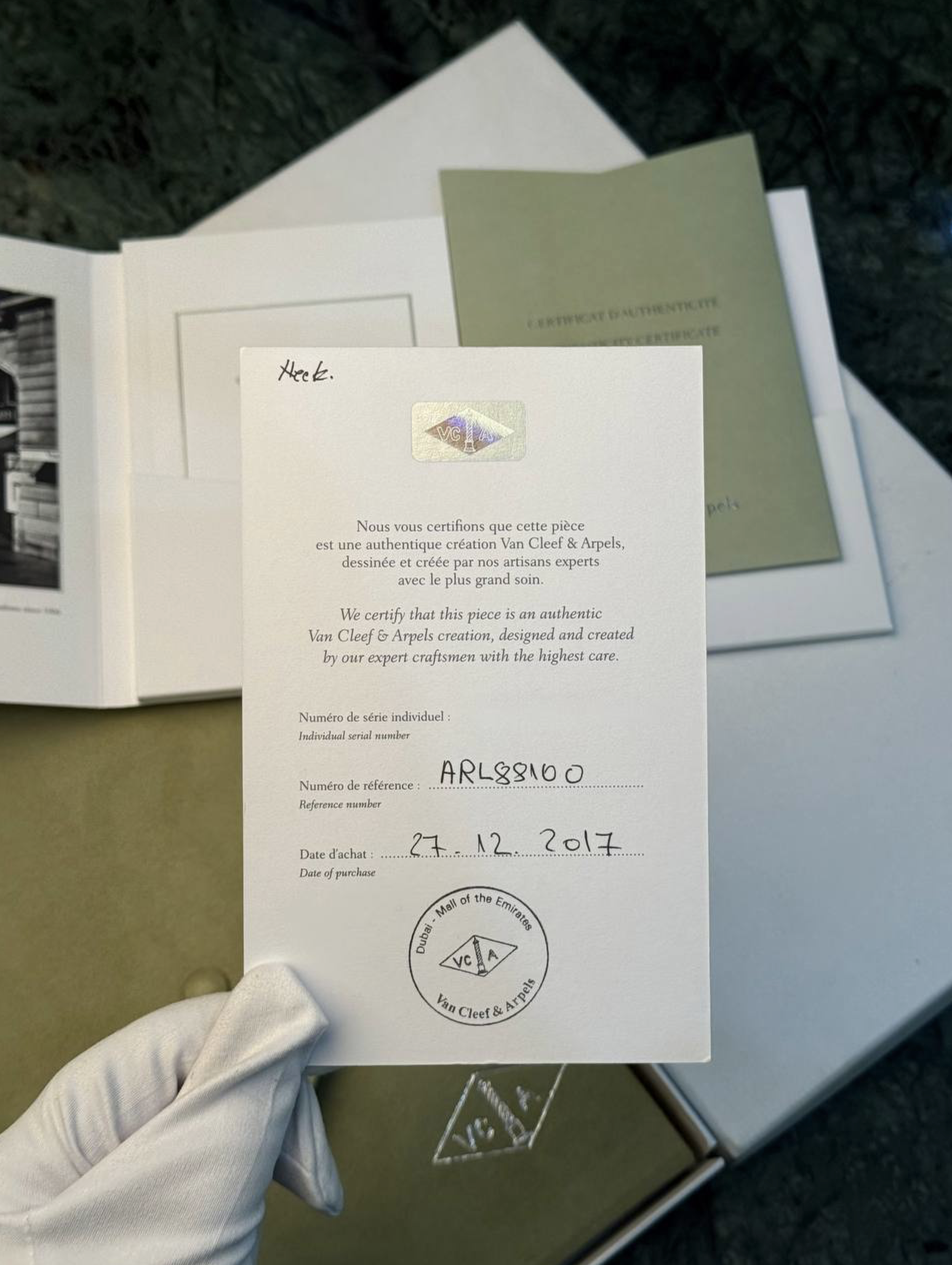 Person wearing a white glove holding an authentication certificate and a Van Cleef & Arpels document with a seal visible. A green booklet rests in the background.
